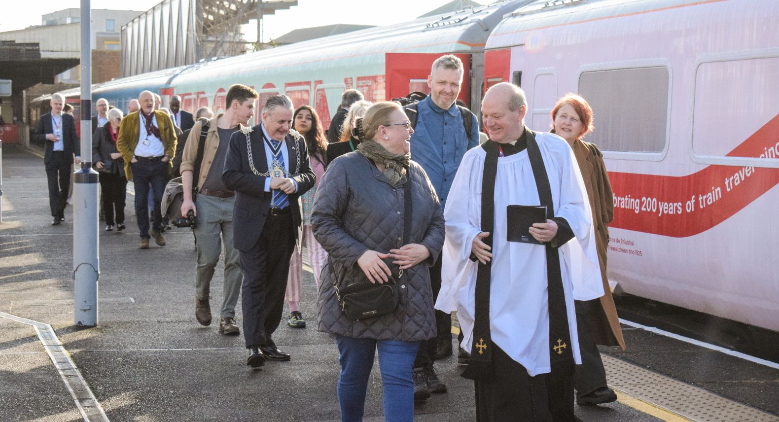 Clergyman leading two columns of people along a platform beside a train