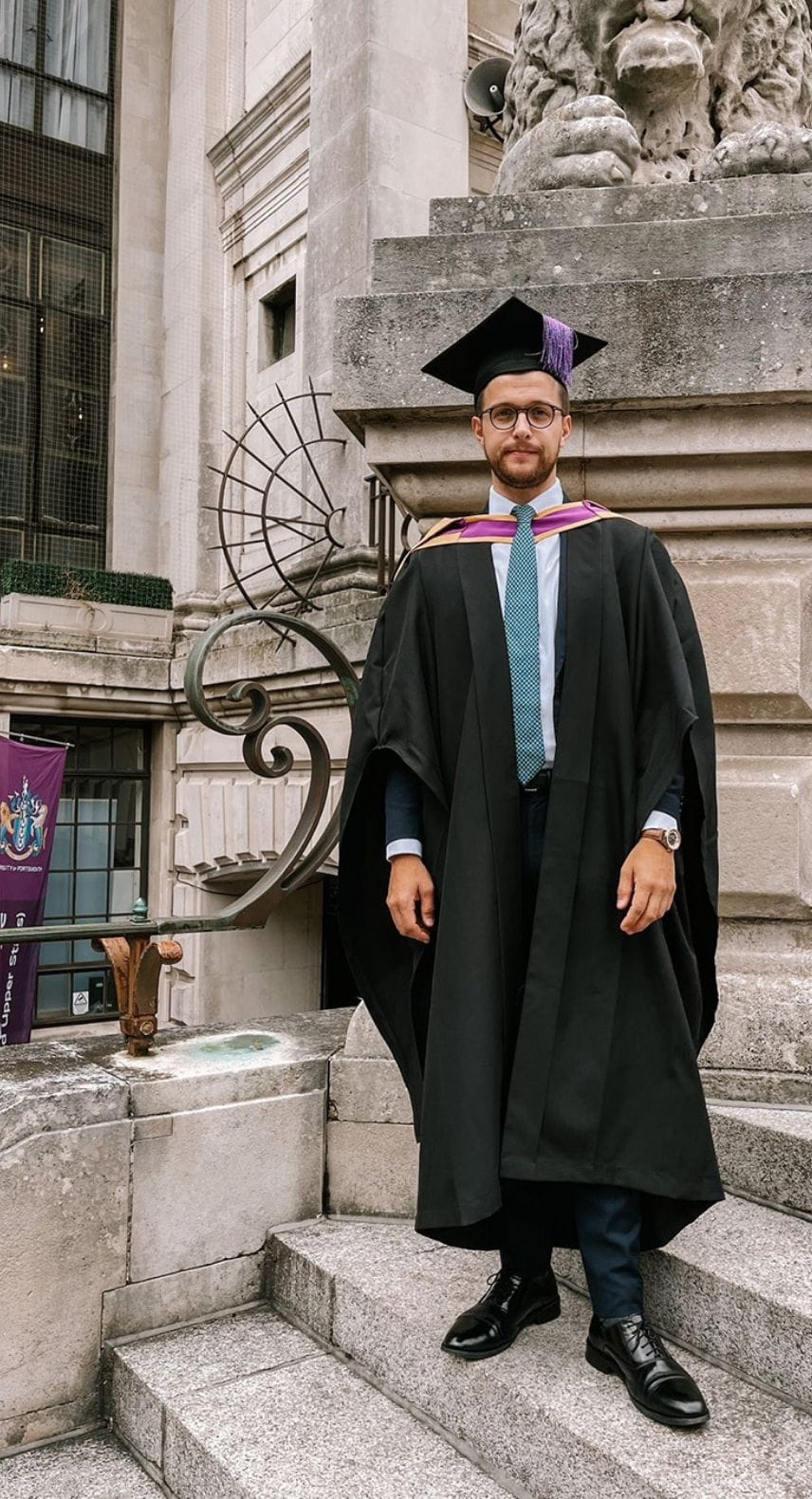 A man in glasses standing on a flight of steps, wearing a mortarboard and gown
