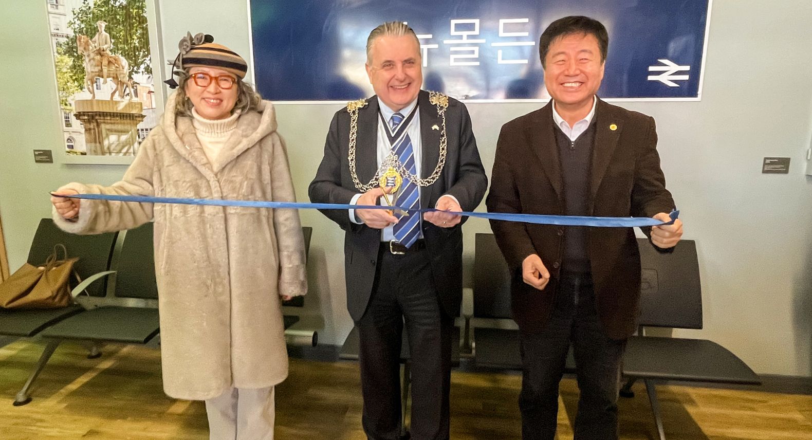 Man wearing a Mayoral chain cutting a ribbon with a Korean man and woman on either side of him