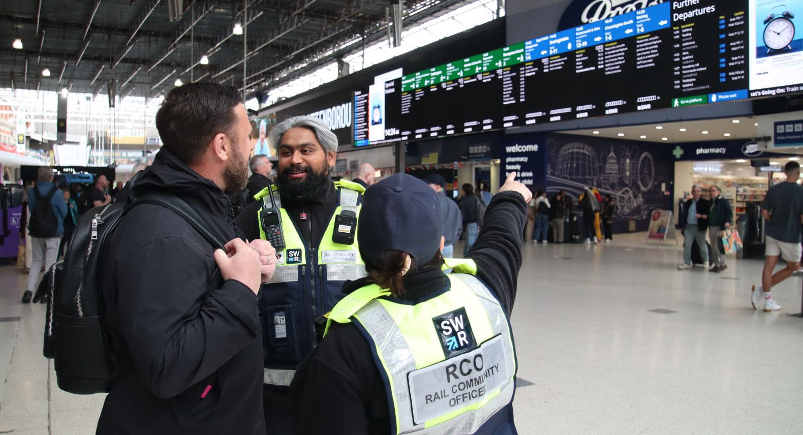 Two Rail Community Officers helping a third person and pointing at the departure board at Waterloo