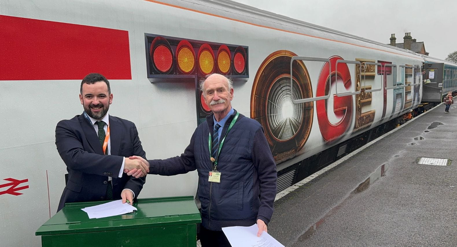Two men standing behind a desk, in front of a train, shaking hands 