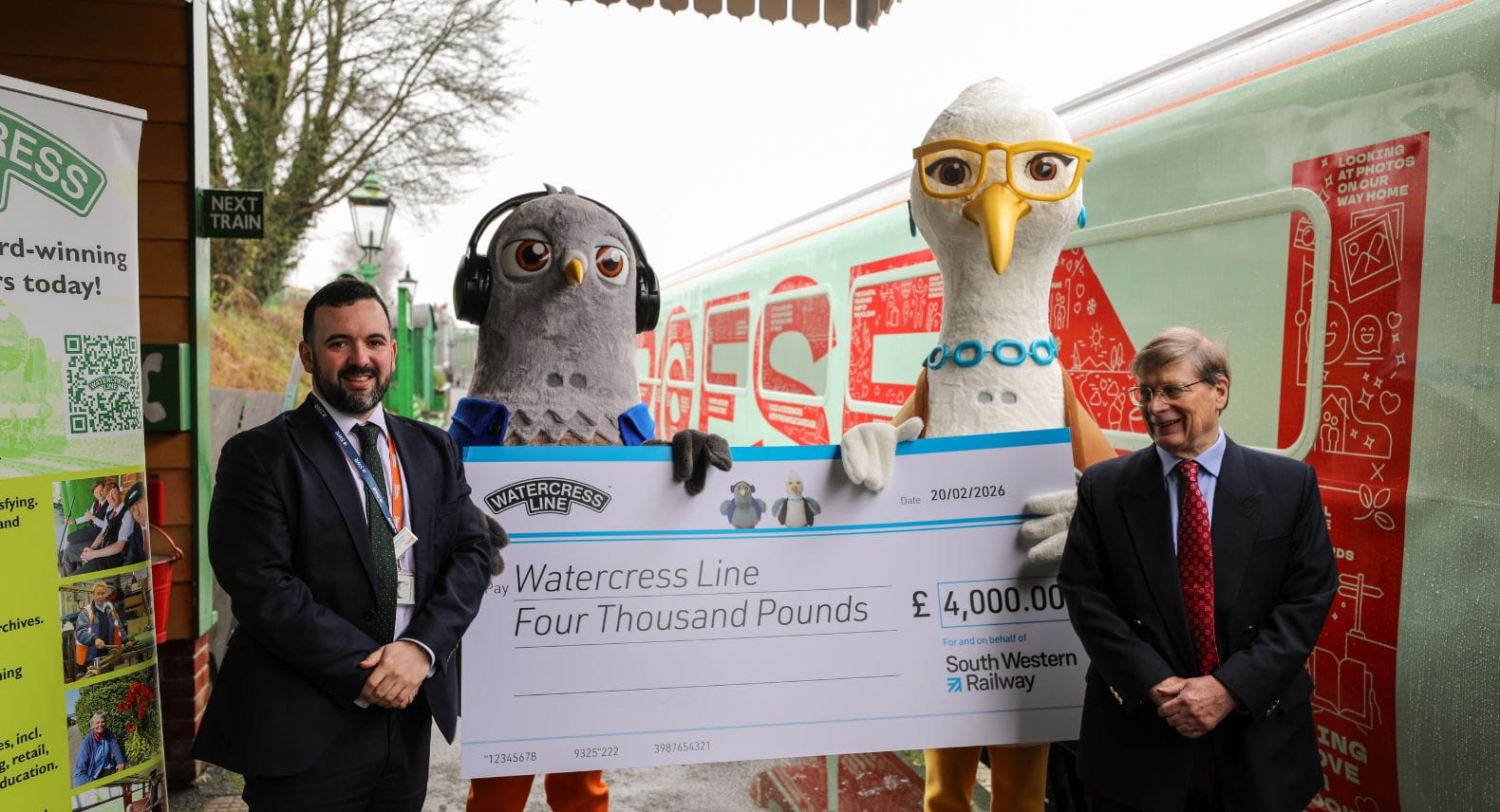 Two men in suits and and a oversized pigeon and seagull mascot holding a giant cheque between them