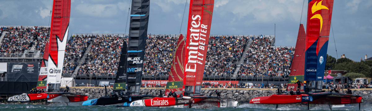 High‑speed catamarans race close to shore during a major sailing competition, each with brightly branded sails including Flyhouse, Black Foils, Emirates, and Oracle. A large crowd fills the grandstands along the rocky waterfront, watching the boats skim across the water on a sunny day.