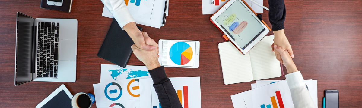 Business meeting with people shaking hands over charts, laptops, and coffee on a wooden table.