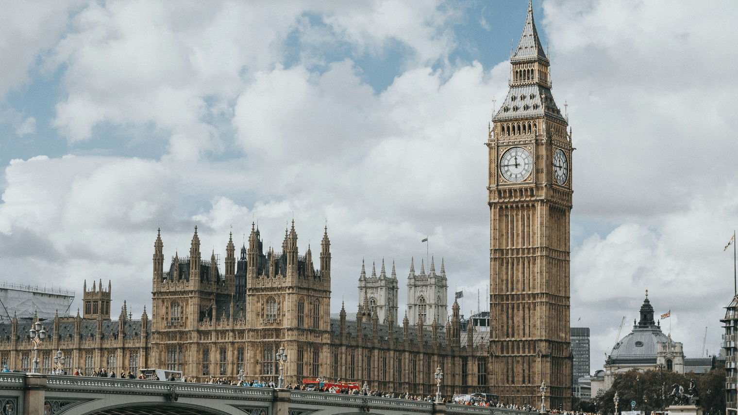 Big Ben and the Palace of Westminster viewed from Westminster Bridge, with pedestrians and a red bus in the foreground.