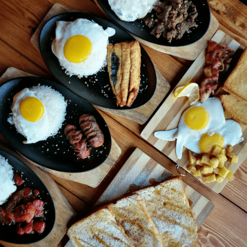 Breakfast plates with rice, sunny‑side‑up eggs, and assorted meats arranged on a wooden table.