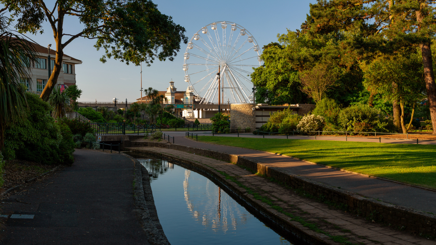 A canal-side path lined with trees, with Bournemouth’s Ferris wheel rising behind buildings and reflecting in the water.