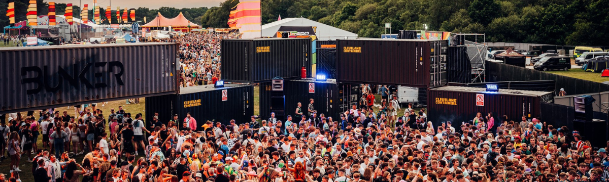 Crowd dancing in a forest under colorful geometric canopy at a psychedelic DJ stage.