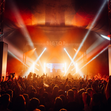 Packed audience inside a tent facing a brightly lit “BIGTOP” concert stage.