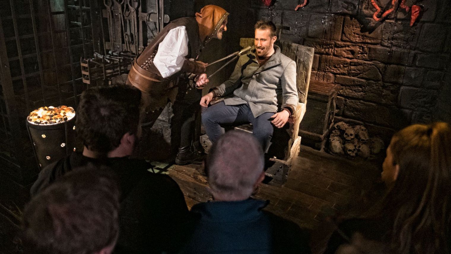 A character in a gloomy lit room in The London Dungeon leans menacingly towards a man dressed in modern clothes who is seated in a chair with his hands strapped to the arms. The character in old Victorian costume is hold a pair of large plyers to the man's mouth while onlookers watch