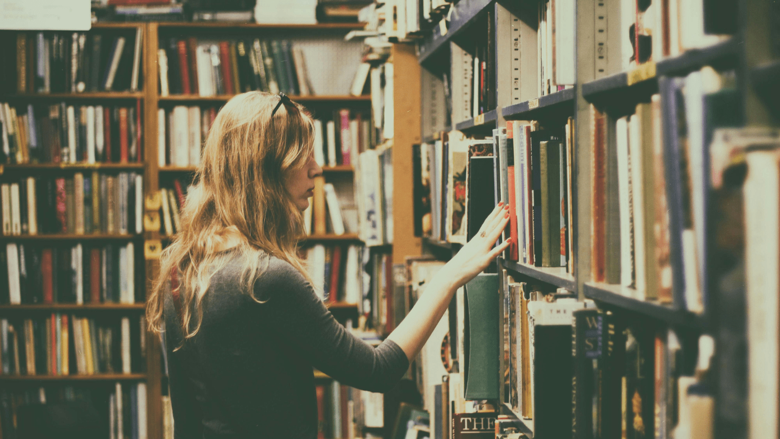 A person stands in front of a full bookshelf in a library or bookstore, extending a hand to pull out a book while browsing the collection.