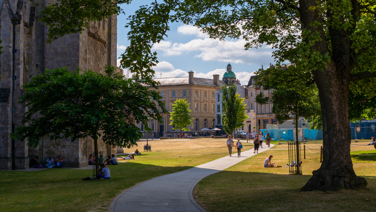 A curved path runs through a sunny park where people sit and walk among historic buildings and trees.