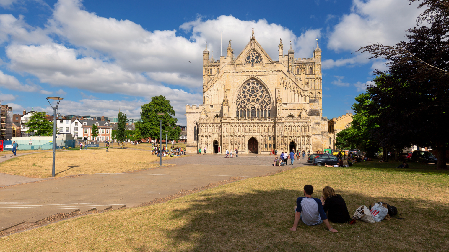 Exeter Cathedral’s ornate Gothic façade overlooks people sitting and walking on the grass in bright weather.