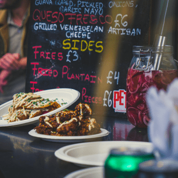 Food stall counter with plates of fried plantain and other dishes in front of a chalkboard menu.